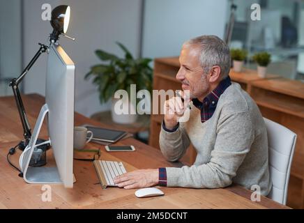 Ich habe ein paar letzte Details zu seinem Projekt gemacht. Ein lächelnder, reifer Geschäftsmann, der an einem Computer in einem Büro arbeitet. Stockfoto