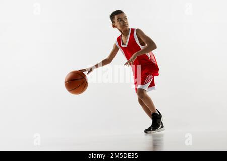 Porträt eines Teenagers in roter Uniform, der Basketball mit grauem Studiohintergrund spielt. Dribbeln Stockfoto