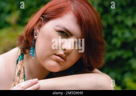 Porträt einer jungen Kaukasier-Latina-Frau argentinischer ethnischer Herkunft, plus Grössenmodell, sitzt draußen, lehnt sich auf ihren Arm, schaut in die Kamera, verschwommen Stockfoto