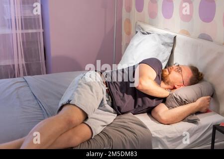 Der Bart eines jungen Mannes schlief in einer Brille ein, auf einem Bett in Kleidung, im Schlafzimmer, mit Licht an. Müdigkeit, Schlafmangel, Stress, Komfort, Zufälligkeit Stockfoto