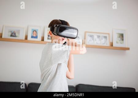 Ein Junge spielt zu Hause Schwertkampf-Videospiele, trägt ein Virtual-Reality-Headset und hält die Fernbedienung in der Hand Stockfoto