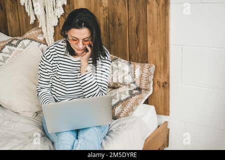 Eine Frau sitzt zu Hause auf dem Bett, telefoniert und schaut in einen Laptop. Stockfoto
