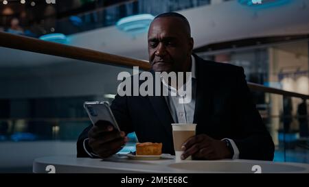 Afrikanischer erwachsener Geschäftsmann mittleren Alters, 50s ethnischer Mann im Anzug Arbeitgeber Unternehmer sitzt am Tisch im Café trinken Kaffee trinken Tee surfen Stockfoto