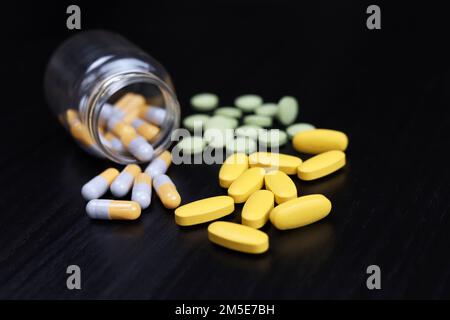 Verschiedene Pillen auf einem schwarzen Holztisch, Variation der Medikamente in Kapseln, die aus einer Flasche verstreut sind. Hintergrund für Pharmazie, Antibiotika, Vitamine Stockfoto