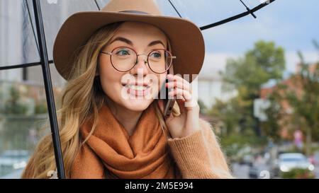 Attraktive junge Frau mit Schirm beantwortet Anrufe auf der Straße im Regen, kluges Mädchen kommuniziert enthusiastisch, emotionale weiße Dame Stockfoto