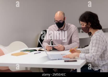 Zwei junge Mitarbeiter in Masken arbeiten zusammen und machen sich Notizen im Büro Stockfoto