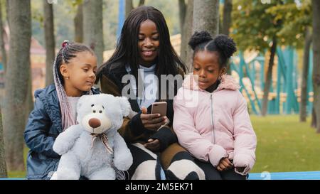 Freundliche Familie, die auf einer Bank im Park sitzt, junge mutter, die auf ihr Handy schaut, Töchter, Kind, das einen Teddybär hält, afroamerikanische Mutter kommuniziert mit ihr Stockfoto