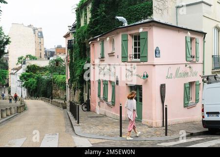 Paris, Frankreich - Frau geht am La Maison Rose Cafe Restaurant in Montmartre vorbei. Hübsches pinkes Haus mit grünen Fensterläden. Charmante Kopfsteinpflasterstraße. Stockfoto