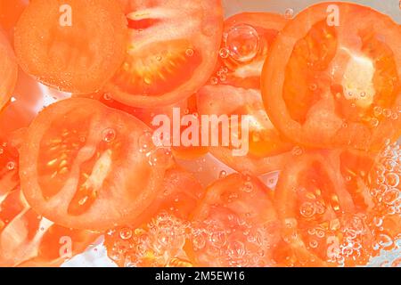 Tomaten-Nahaufnahme in Flüssigkeit mit Blasen. Scheiben roter reifer Tomaten im Wasser. Nahaufnahme frischer Scheiben roter Tomaten auf weißem Hintergrund Stockfoto