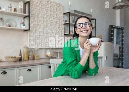 Junge schöne Frau, Mutter trinkt Kaffee, Tee allein in der Küche zu Hause. Er hält eine Tasse in der Hand, genießt, ruht sich aus, lächelt vor die Kamera. Stockfoto