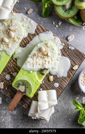 Hausgemachtes veganes Kiwieis oder Eis am Stiel mit weißer Schokolade und Nüssen auf Holzbrett, Blick von oben, flach liegend. Vertikal Stockfoto
