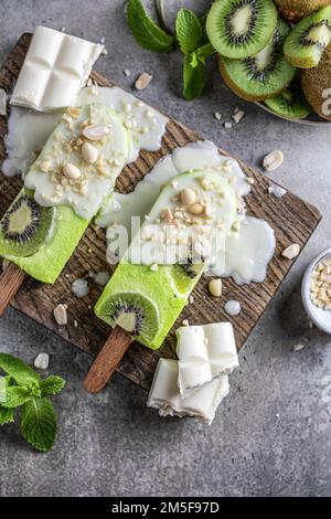 Hausgemachtes veganes Kiwieis oder Eis am Stiel mit weißer Schokolade und Nüssen auf Holzbrett, Blick von oben, flach liegend. Vertikal Stockfoto