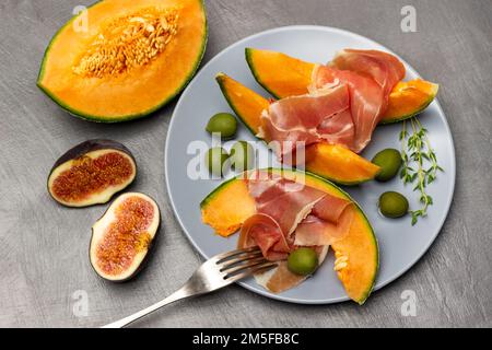 Jamon auf einer Scheibe Melone, grüne Oliven und Thymianzweige auf grauen Tellern. Zwei Hälften Feigen und Melone auf dem Tisch. Draufsicht. Grauer Hintergrund. Stockfoto