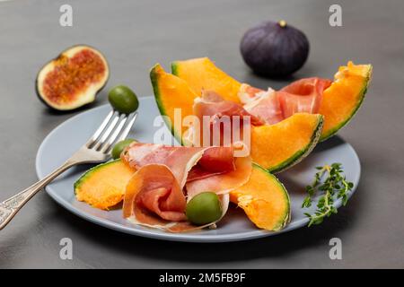 Grüne Oliven auf einer Gabel, Jamon auf einer Melonenscheibe, Thymianzweige auf einem grauen Teller. Frische Feigen auf dem Tisch. Stockfoto