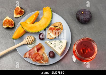 Honigstäbchen und Käse, braune Oliven, Jamon und Melone auf grauem Teller. Ein Glas Wein und eine Hälfte Feigen auf dem Tisch. Flach verlegt. Grauer Hintergrund. Stockfoto
