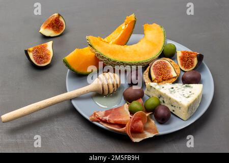 Braune und grüne Oliven, Honigstäbchen und Käse, Jamon und Melone auf grauem Teller. Draufsicht. Grauer Hintergrund. Stockfoto