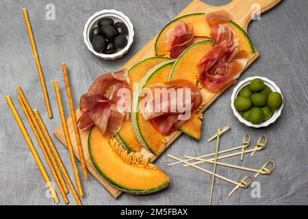 Schwarze und grüne Oliven in Schüsseln. Jamon auf einem Stück Melone auf dem Schneidebrett. Flach verlegt. Grauer Hintergrund. Stockfoto