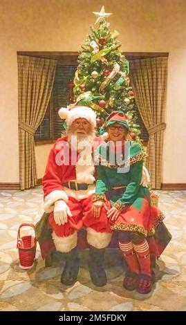 Der Weihnachtsmann und seine Elfe sitzen vor dem Weihnachtsbaum und begrüßen die Kinder an einem Winterabend in Marine auf St. Croix, Minnesota, USA. Stockfoto