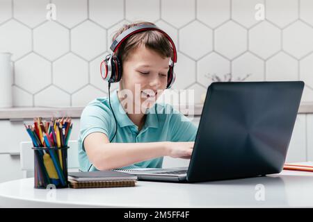 Porträt eines jungen, lächelnden Jungen mit T-Shirt, Kopfhörern, am Schreibtisch neben Bleistiften sitzen, Laptop benutzen, tippen, Spiele spielen, Nachrichten senden Stockfoto