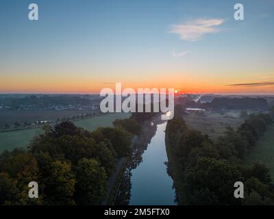 Von einer Drohne aus der Vogelperspektive sehen Sie den Sonnenaufgang oder -Untergang hinter dem Fluss und füllen die Bäume mit warmem Abendlicht, der Nebel steigt in den goldenen Lichtstrahlen auf. Wolken und Wolken werden im Wasser reflektiert. Hochwertiges Foto Stockfoto