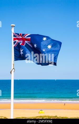 Australische Flagge, die an einem sonnigen Tag gegen eine Strandszene fliegt, NSW Australien. Stockfoto