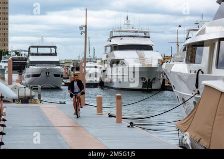 Ein Mann fährt mit dem Fahrrad auf dem Pier inmitten der Motoryachten, die an der Palm Harbor Marina an der Lake Worth Lagoon in West Palm Beach, Florida, USA angelegt sind. Stockfoto