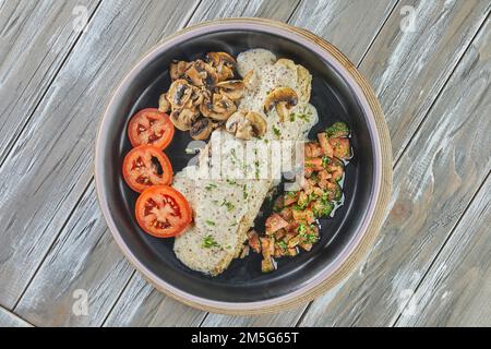 Wolfsbarsch mit Senf, Pilzen und Tomaten auf schwarzem Teller. Flach verlegt. Stockfoto