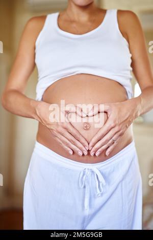 Bedingungslose Liebe. Eine schwangere Frau, die ihren Bauch mit ihren Händen hält und eine Herzform bildet. Stockfoto