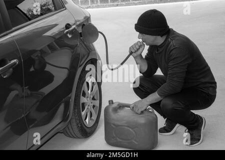 Bild eines schwarz gekleideten Diebes, der mit einem Gummischlauch Benzin aus dem Tank saugt. Hinweis auf die Zunahme des Kraftstoffdiebstahls aufgrund überhöhter Kosten. Stockfoto