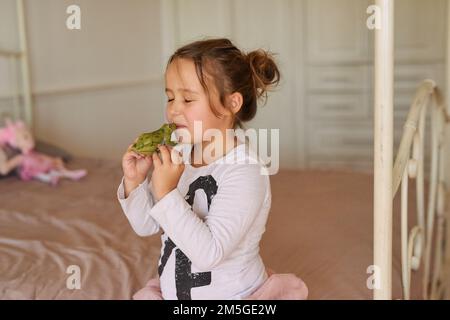 Vielleicht verwandelt es sich in einen kleinen Prinzen. Ein kleines Mädchen, das zu Hause einen Spielzeugfrosch küsst. Stockfoto