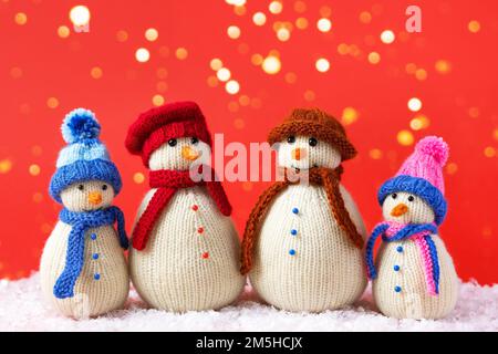 Eine Familie gestrickter Schneemänner feiert die Feiertage auf rotem Hintergrund mit goldenen Lichtern. Wintermärchen-Geschichte mit eigenen Händen, Hobby. Merry Chr Stockfoto
