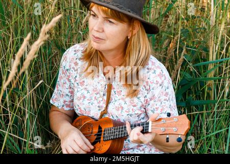 Unschärfe-Frau mit Gitarre auf Naturhintergrund. Frauen draußen auf der grünen Wiese. Ukulele-Akkord. Sommerzeit. Ein Volkskünstler. Lächelnde Modefrau. Raus Stockfoto