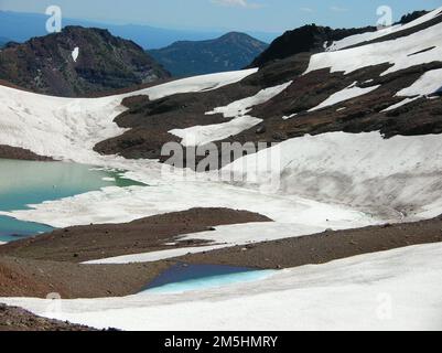 Cascade Lakes Scenic Byway - Sky Blue Waters und schneebedeckte Pisten. Sanfte Hänge führen hinunter in ruhiges blaues Wasser, umgeben von Felsen, Erde und Schnee. Standort: Oregon (44,122° N 121,770° W) Stockfoto