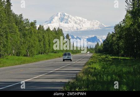 Der George Parks Highway Scenic Byway – Fahrt entlang des George Parks Highway. Die Reisenden fahren entlang der Autobahn in Richtung schneebedeckter Berge. Grüne Bäume säumen die Seiten der Nebenstraße. Standort: Alaska Stockfoto