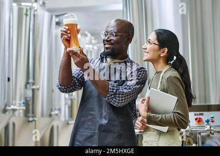 Ein afroamerikanischer Ingenieur untersuchte frisches Bier in Glas und bespricht die Qualität mit seinem Kollegen Stockfoto