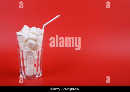 Glas mit Strohhalm gefüllt mit Zuckerwürfeln, isoliert auf rotem Hintergrund. Eine Metapher für die Tatsache, dass Getränke oft viel Zucker enthalten Stockfoto