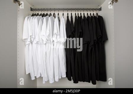 Schwarz-weiße T-Shirts hängen an einer Kleiderstange in einem weißen Kleiderschrank Stockfoto