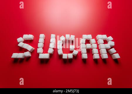 Das Wort Zucker besteht aus Zuckerwürfeln auf rotem Hintergrund. Foto zum Thema Gefahren des Verzehrs von raffiniertem Zucker Stockfoto
