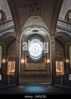 Louvre Palast symmetrische architektonische Details einer Halle mit Steintreppe, kunstvollen Geländern, verschiedenen Korridoren und einem Uhrenfenster in der Mitte, Stockfoto