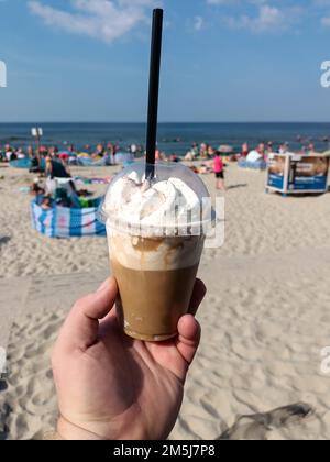 Eine Hand mit einer Plastiktasse gefrorenen Kaffees am Strand. Stockfoto