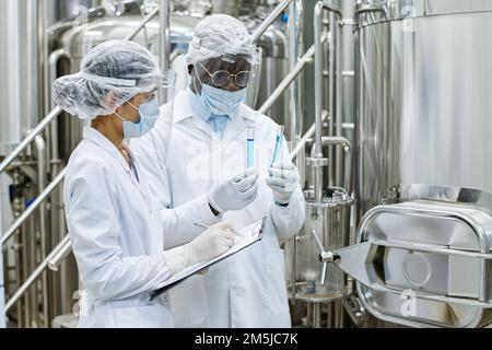 Afroamerikanischer Wissenschaftler, der Flüssigkeit in Reagenzgläsern untersucht, während sein Assistent während eines wissenschaftlichen Experiments Notizen im Dokument macht Stockfoto