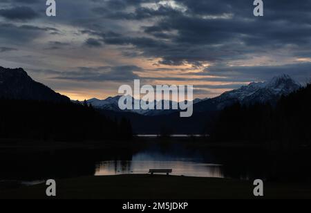 29. Dezember 2022, Bayern, Roßhaupten: Eine Bank steht im Sonnenuntergang am Ufer des Illasberger Sees vor den Alpen, die im Sonnenuntergang liegen. Foto: Karl-Josef Hildenbrand/dpa Stockfoto