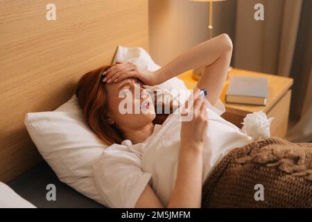 Krankheitsmüde Frau, die die Stirn berührt, nachdem sie ein Thermometer benutzt hat, um die Körpertemperatur zu überprüfen und Medikamente unter der Bettdecke zu nehmen. Stockfoto