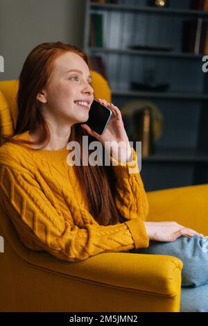 Vertikales Porträt einer fröhlichen jungen Rothaarigen, die zu Hause auf einem gelben Stuhl sitzt und mit einem Smartphone spricht. Hübsche Dame, die sich lustig macht Stockfoto