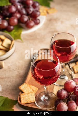 Zwei Gläser Rotwein, ein Haufen rote Trauben, Cracker und Korkenzieher auf einem Holztablett mit beigefarbenem strukturiertem Hintergrund. Feier für zwei Konzerte Stockfoto
