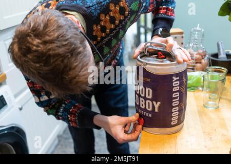 Ein Mann zu Hause, der einen Weihnachtspullover trägt, der den Wasserhahn auf einem Mini-Fass von Good Old Boy Bitter öffnet, um ein Bier zu Weihnachten in England einzuschenken Stockfoto