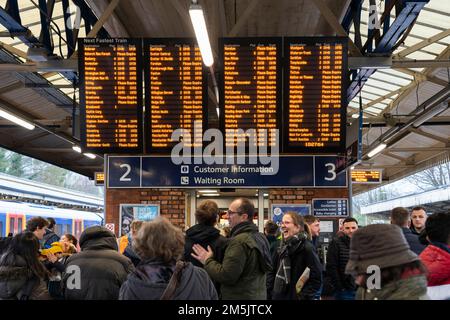 Pendler und Bahnpassagiere am Bahnhof Basingstoke stehen und warten auf einem Bahnsteig unter einer Abfahrtafel, die die Zugzeiten anzeigt. England Stockfoto