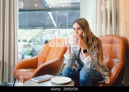 Junge Frau sitzt auf einem Sessel, trinkt Kaffee an einem modernen, offenen Arbeitsplatz, wartet auf einen späten Freund. Stockfoto