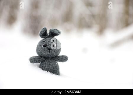 Graues gestricktes Kaninchen im Schnee auf verschwommenem Waldhintergrund, Grußkarte. Symbol des chinesischen Neujahrs 2023 Stockfoto