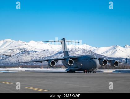 EIN US-AMERIKANISCHER Air Force C-17 Globemaster III, dem 62. Luftwaffenflügel zugeteilt, Joint Base Lewis-McChord, Washington, Taxis auf einer Landebahn, während ein F-22 Raptor, der dem 3. Flügel zugeteilt wurde, während der Übung Rainer war 22A von der Fluglinie abhebt, auf der Joint Base Elmendorf-Richardson, Alaska, 21. März 2022. Rainier war ist eine halbjährliche, große Bereitschaftsübung unter der Leitung des 62. Airlift Wing. Es soll Flugbesatzungen nach realistischen Szenarien Schulen, die ein vollständiges Spektrum an Einsatzbereitschaft gegen moderne Bedrohungen unterstützen und den heutigen Notfallbetrieb nachbilden. Stockfoto